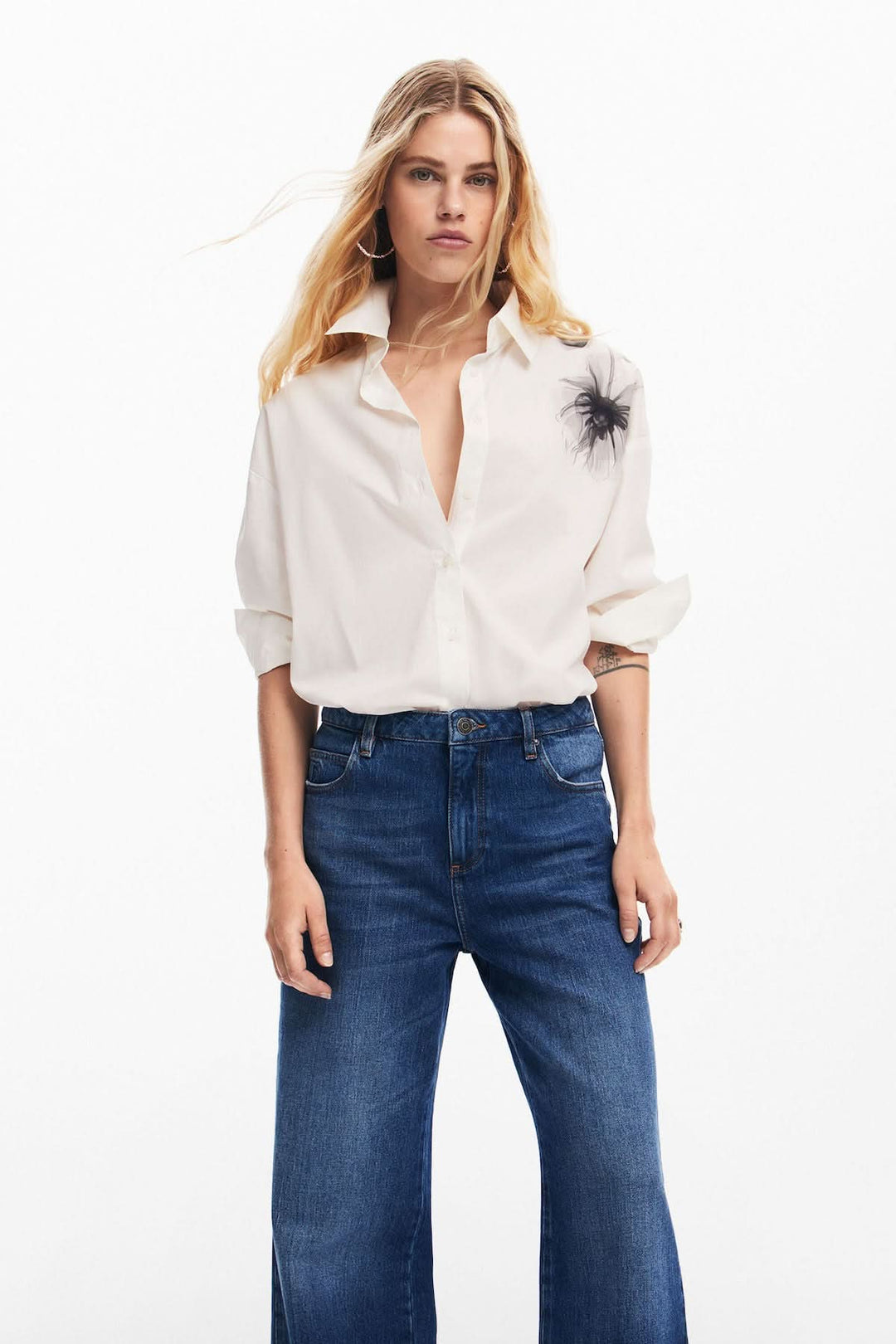 Femme portant une chemise blanche à col et un jean ample, avec un motif floral sur l'épaule.