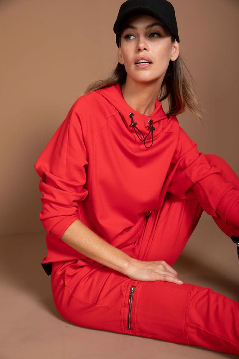 Femme portant un jogging ajusté rouge avec détails utilitaires, style moderne et sportif chic, assise sur un fond beige.