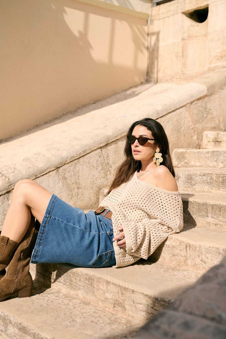 Femme portant une jupe en jean et un pull décontracté, assise sur des escaliers ensoleillés.