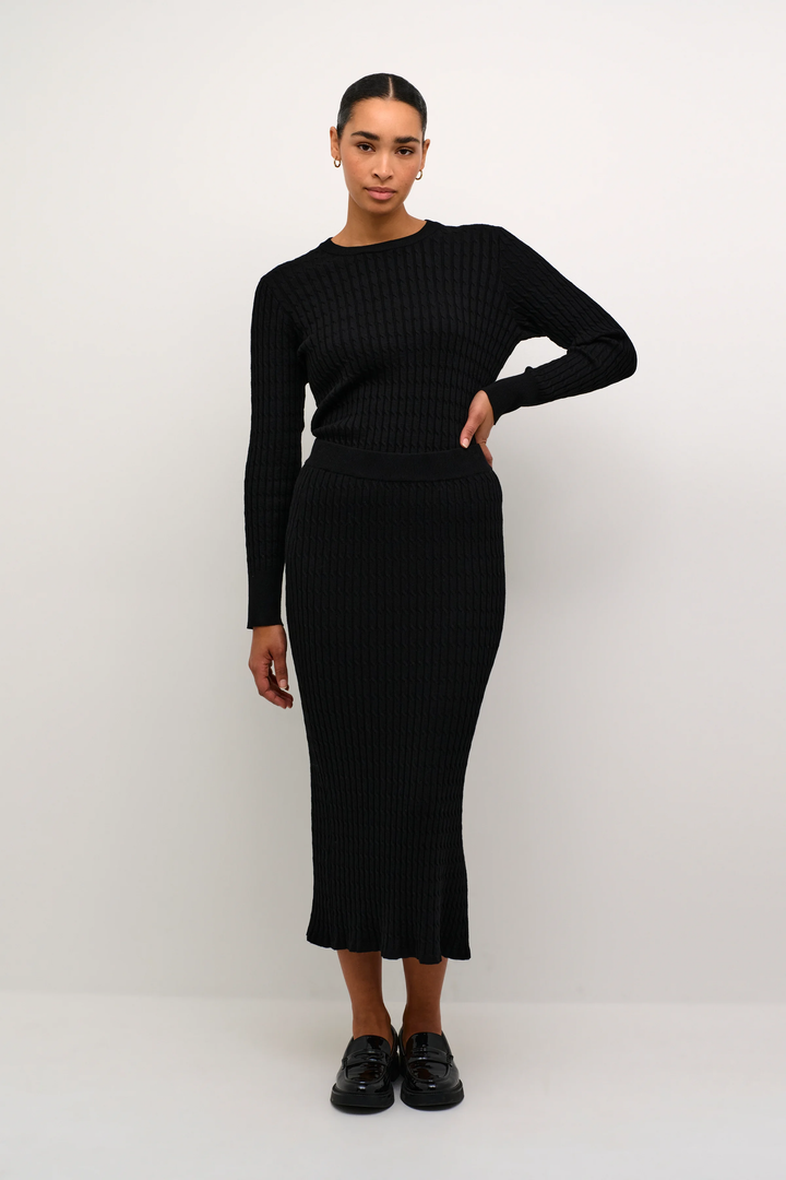 Femme portant une jupe longue noire à fente et un haut assorti, offrant un look élégant et moderne.