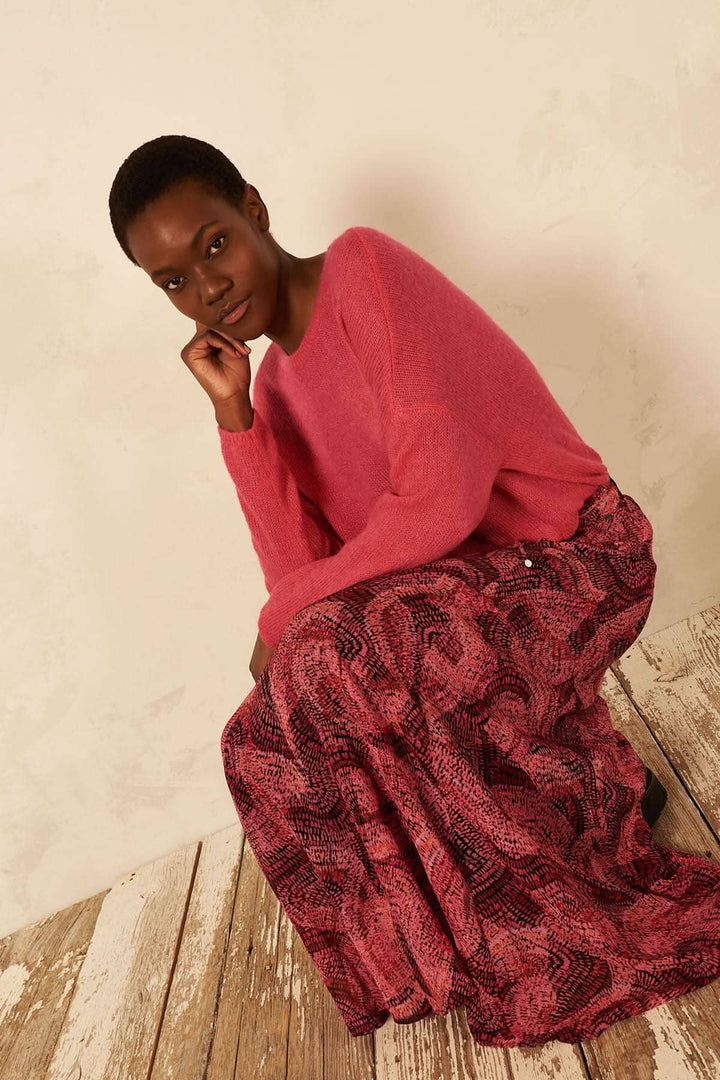 Femme assise en jupe longue abstraite avec un pull rose, sur un sol en bois, en pose contemplative.