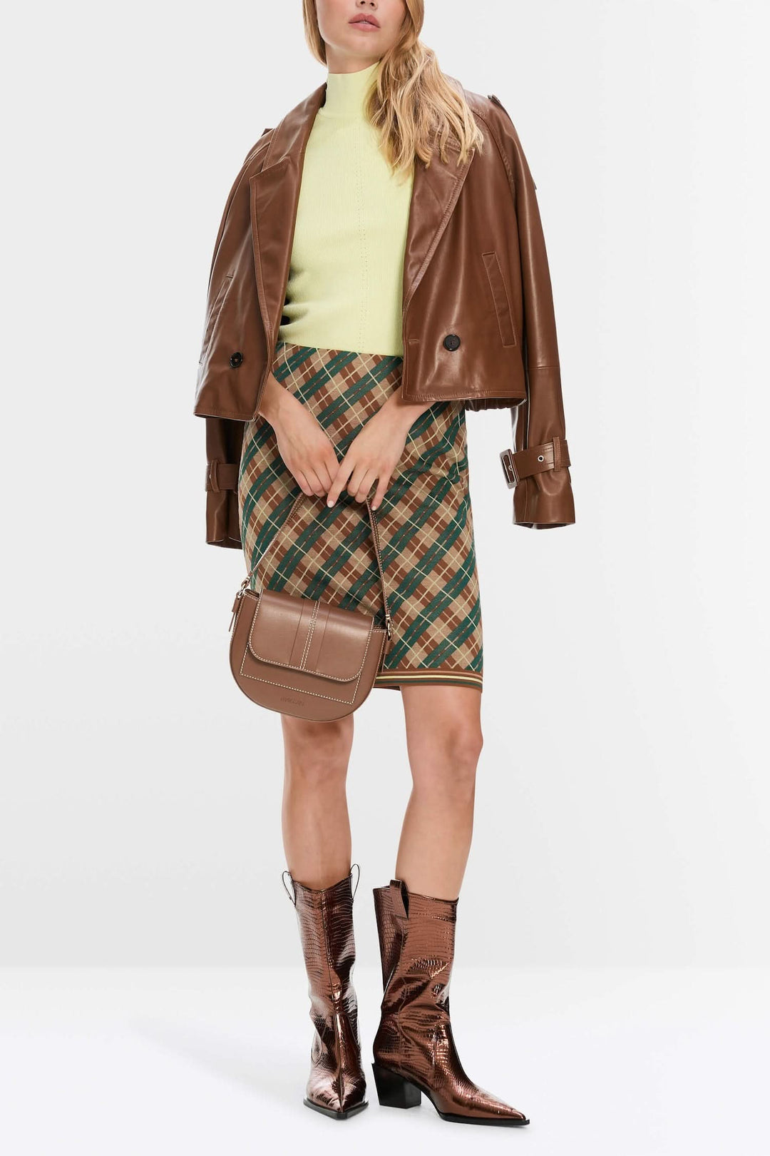 Modèle portant une mini-jupe à carreaux, un pull vert et une veste en cuir, avec des bottes brillantes.