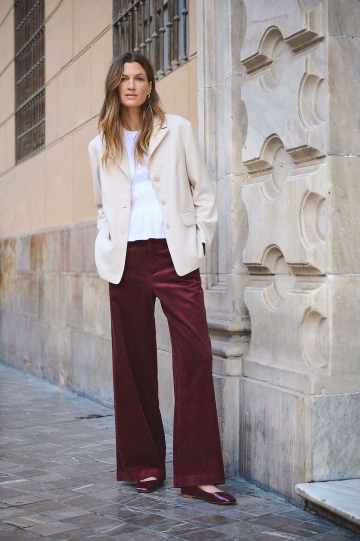 Femme en pantalon velours côtelé bordeaux, veste beige, sur la rue avec une allure rétro-chic.