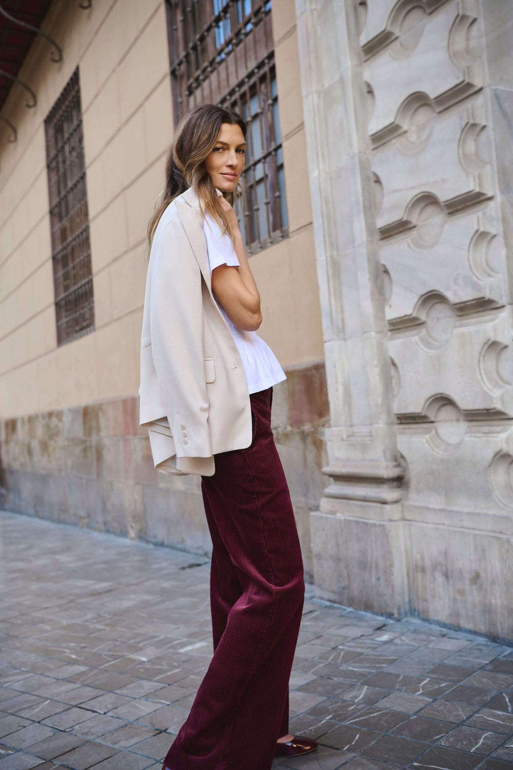 Femme porte un pantalon en velours côtelé rouge et un blazer beige, look élégant et rétro-chic, rue en arrière-plan.