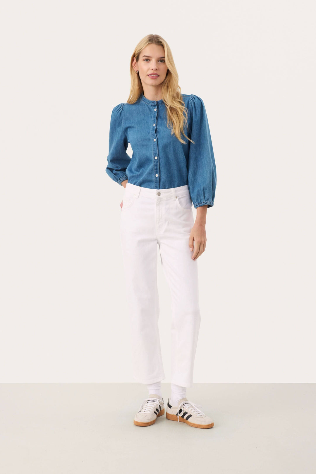 Femme en chemise denim bleu et pantalon blanc, look moderne et décontracté avec des baskets.