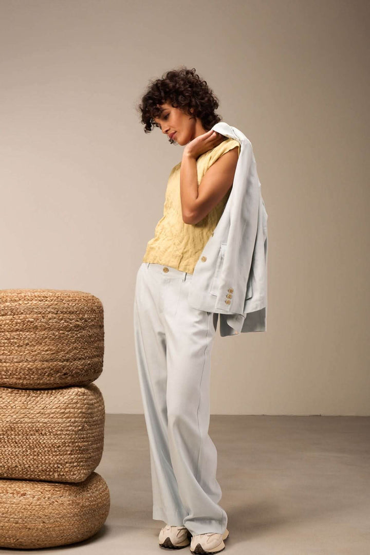 Femme portant un pantalon large bleu clair avec un haut jaune et une veste, se tenant près de poufs.