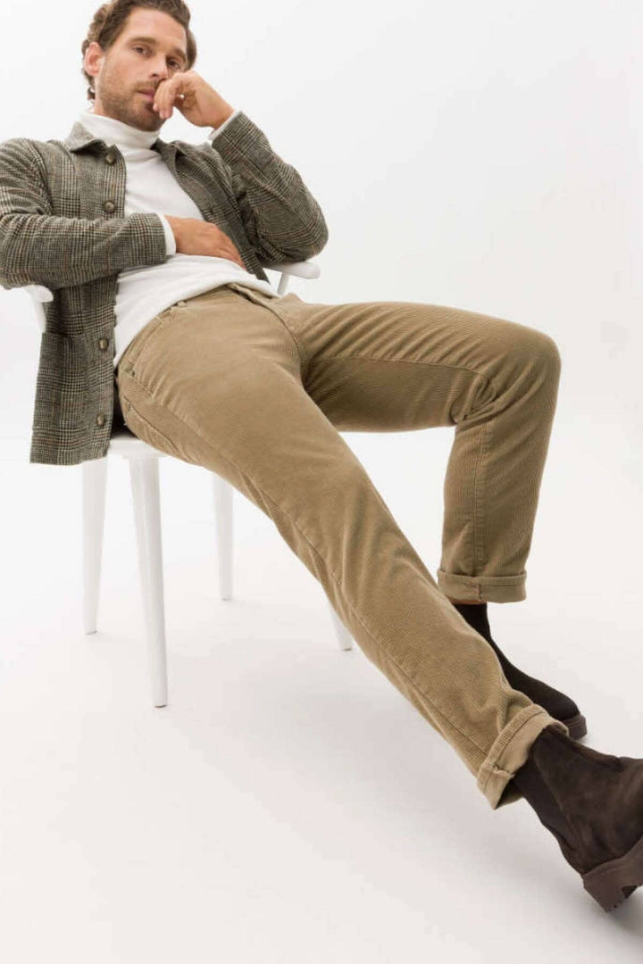 Homme portant un pantalon en corduroy beige et une chemise à carreaux, assis sur une chaise blanche.