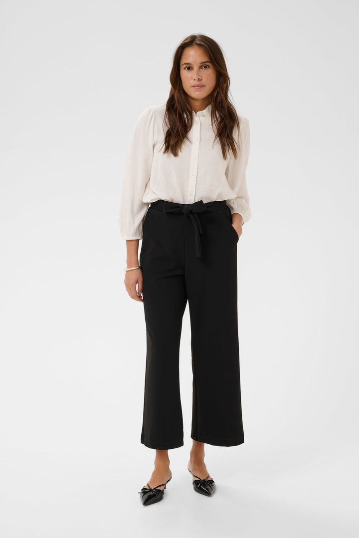 Femme portant un Pantalon jambe large Phoebe noir avec une blouse blanche, élégant et confortable pour un look bureau ou week-end.