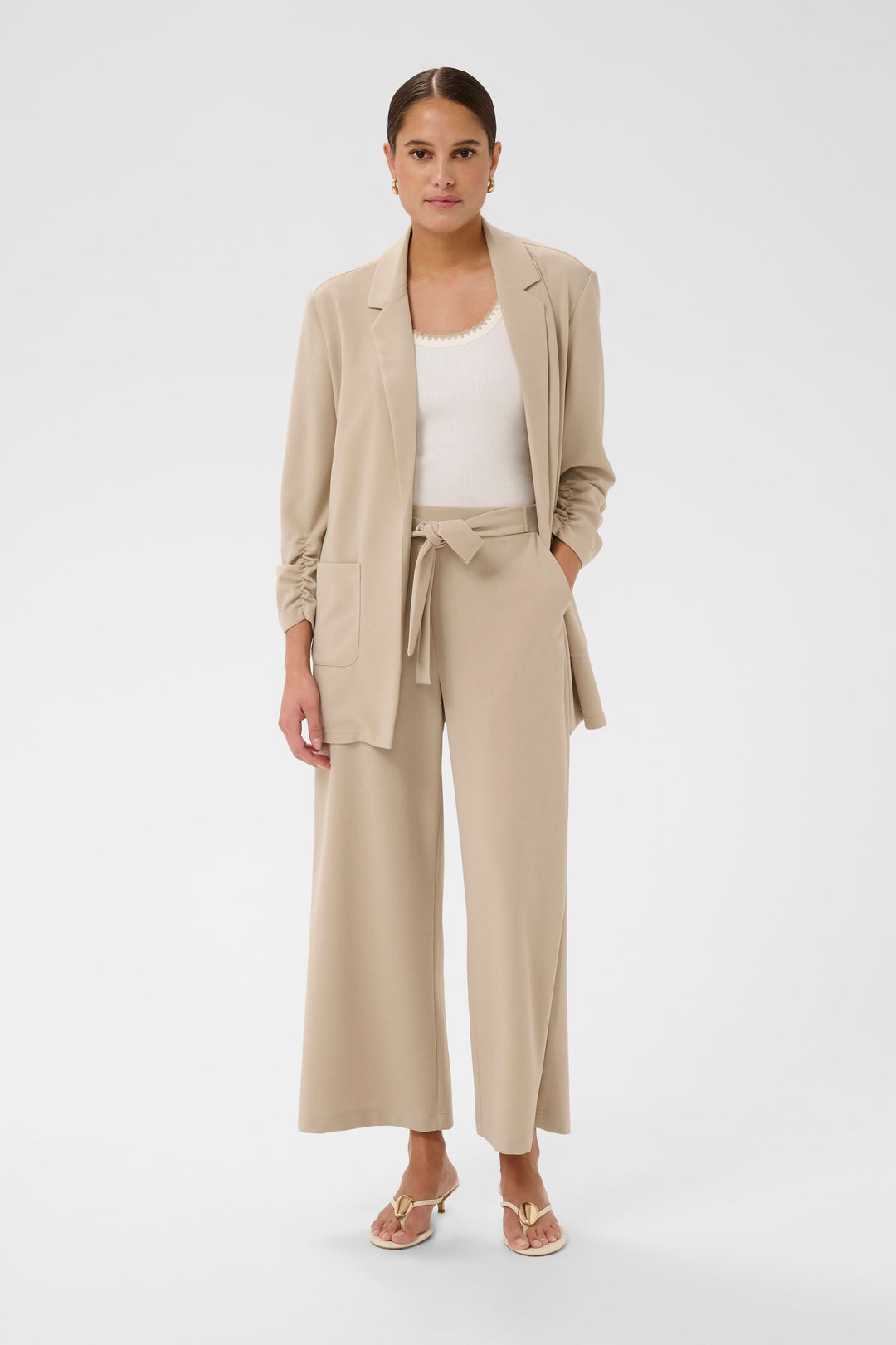 Femme portant un Pantalon jambe large Phoebe beige avec une ceinture nouée, accompagné d'une veste et sandales élégantes.
