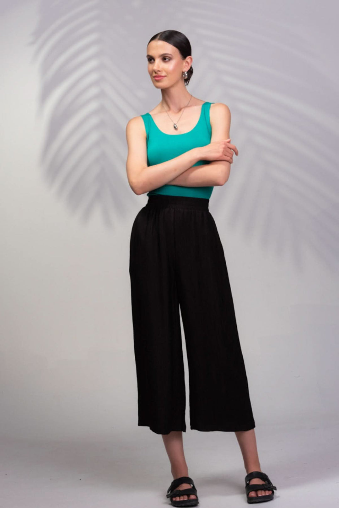 Femme portant un pantalon texturé noir et un débardeur vert, se tenant debout avec un fond de feuille.