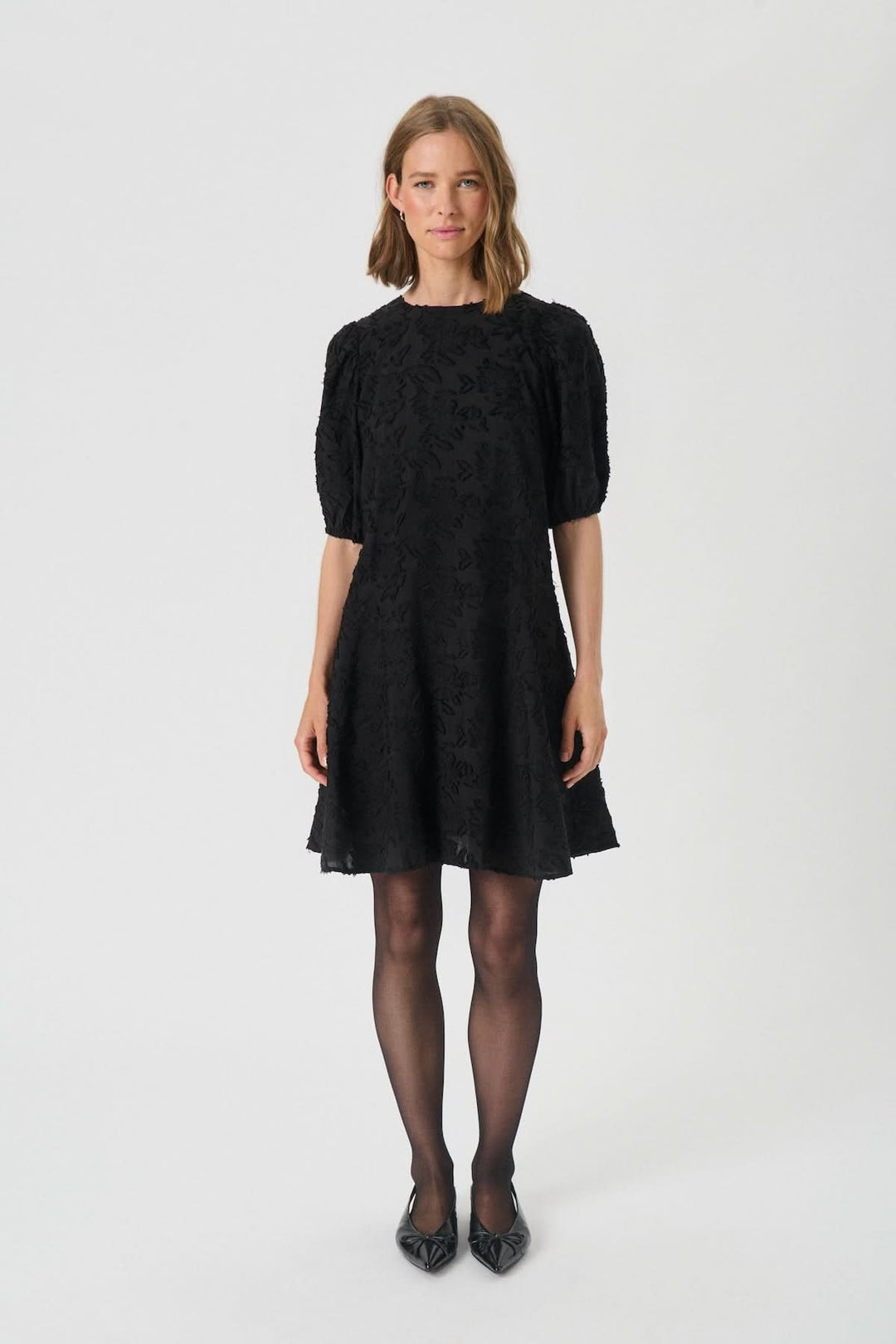 Femme portant la Robe Vilhelmina en dentelle à manches bouffantes, texture florale et coupe évasée, idéale pour événements chic.