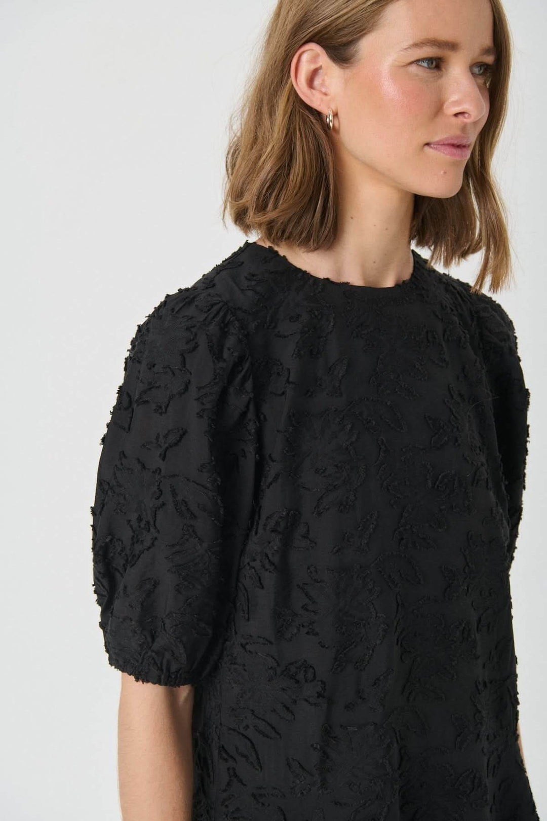 Femme portant la Robe Vilhelmina en dentelle à manches bouffantes, élégante et féminine avec texture florale ton sur ton.