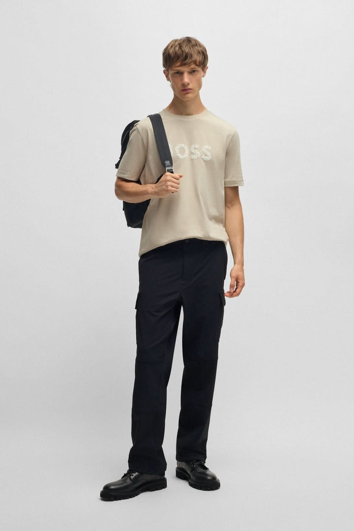 Homme en t-shirt de coton beige avec logo texturé, portant un sac à dos, style décontracté et moderne.