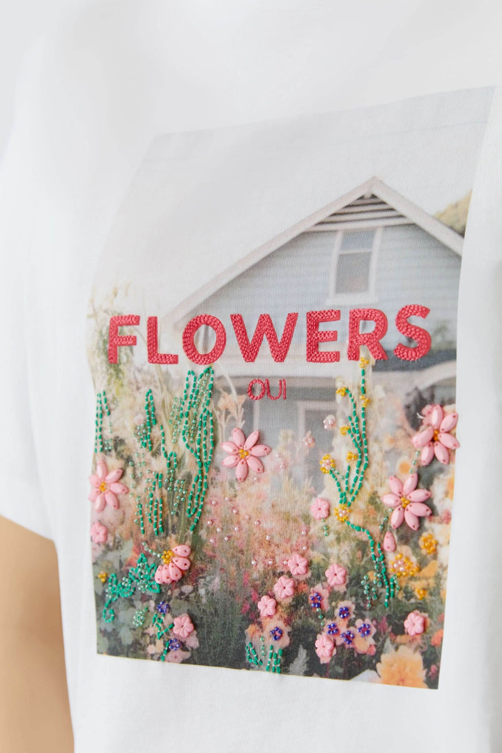T-shirt imprimé graphique avec design floral et texte "FLOWERS oui" sur fond de maison.