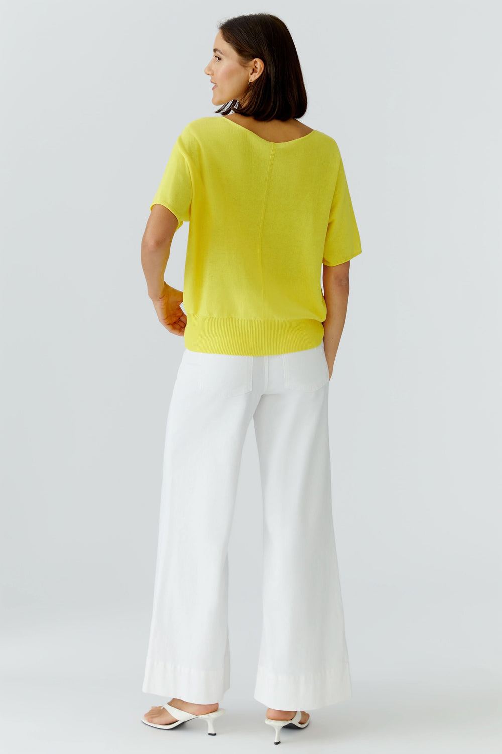 Femme portant un tricot fluide à manches courtes jaune, coupe fluide avec poche poitrine, associé à un pantalon blanc ample et des talons.