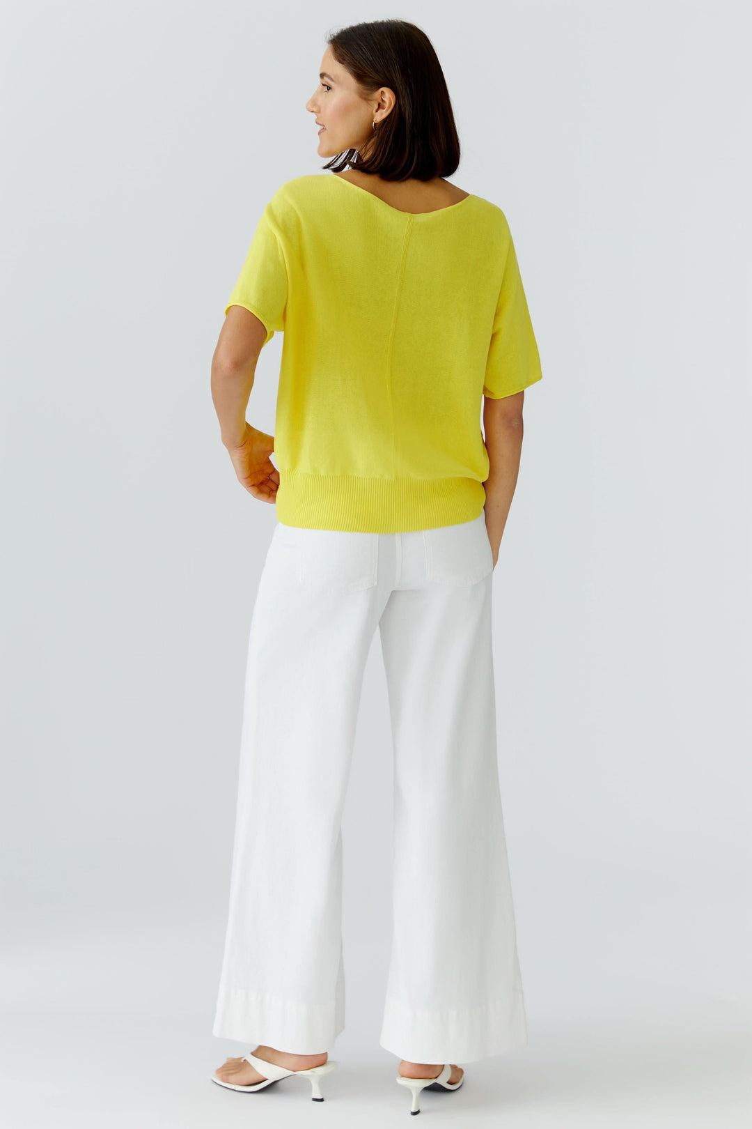 Femme portant un tricot fluide à manches courtes jaune, coupe fluide avec poche poitrine, associé à un pantalon blanc ample et des talons.