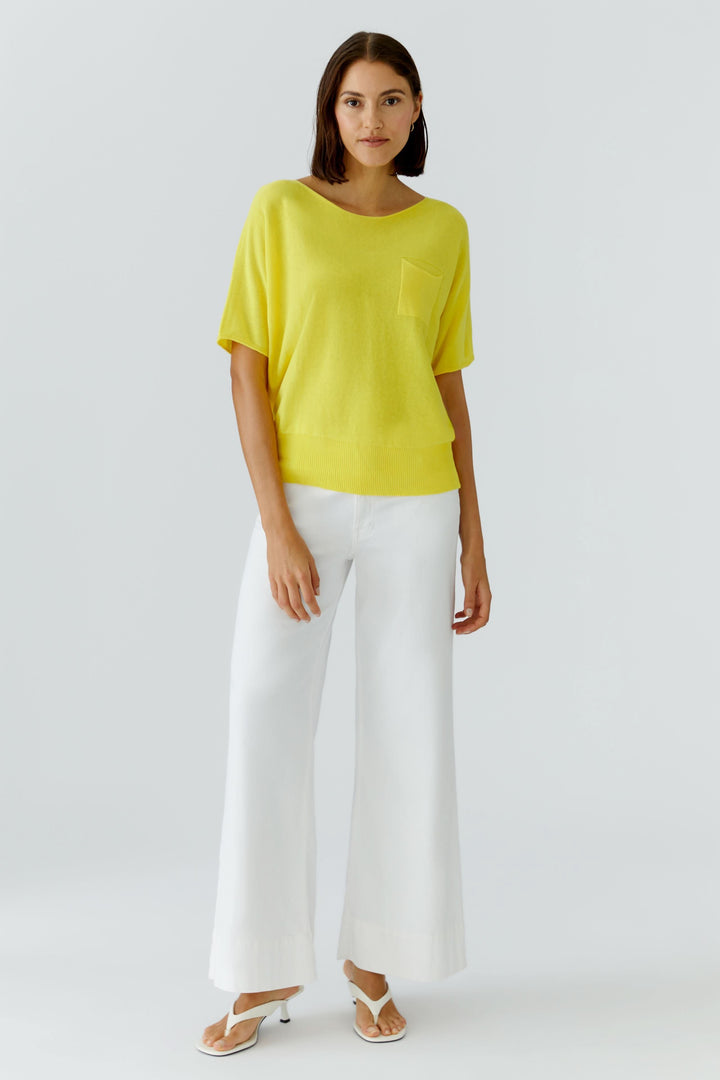 Femme portant un tricot fluide à manches courtes jaune avec col dégagé et poche poitrine, style décontracté et moderne.