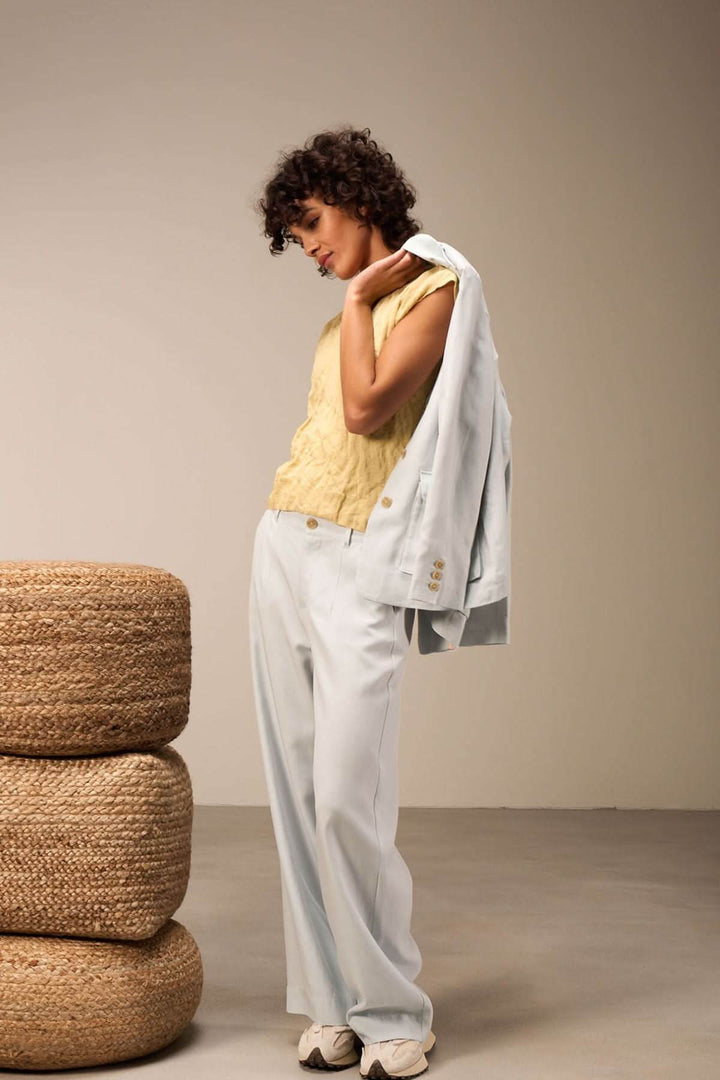 Femme en haut jaune et pantalon bleu clair, portant une veste sur l'épaule, dans un décor minimaliste.