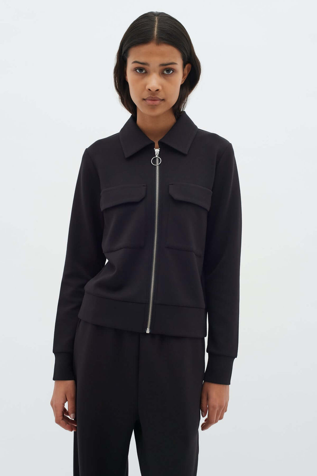 Femme portant une Veste courte zippée Beca noire avec fermeture éclair et poches, style moderne et structuré.