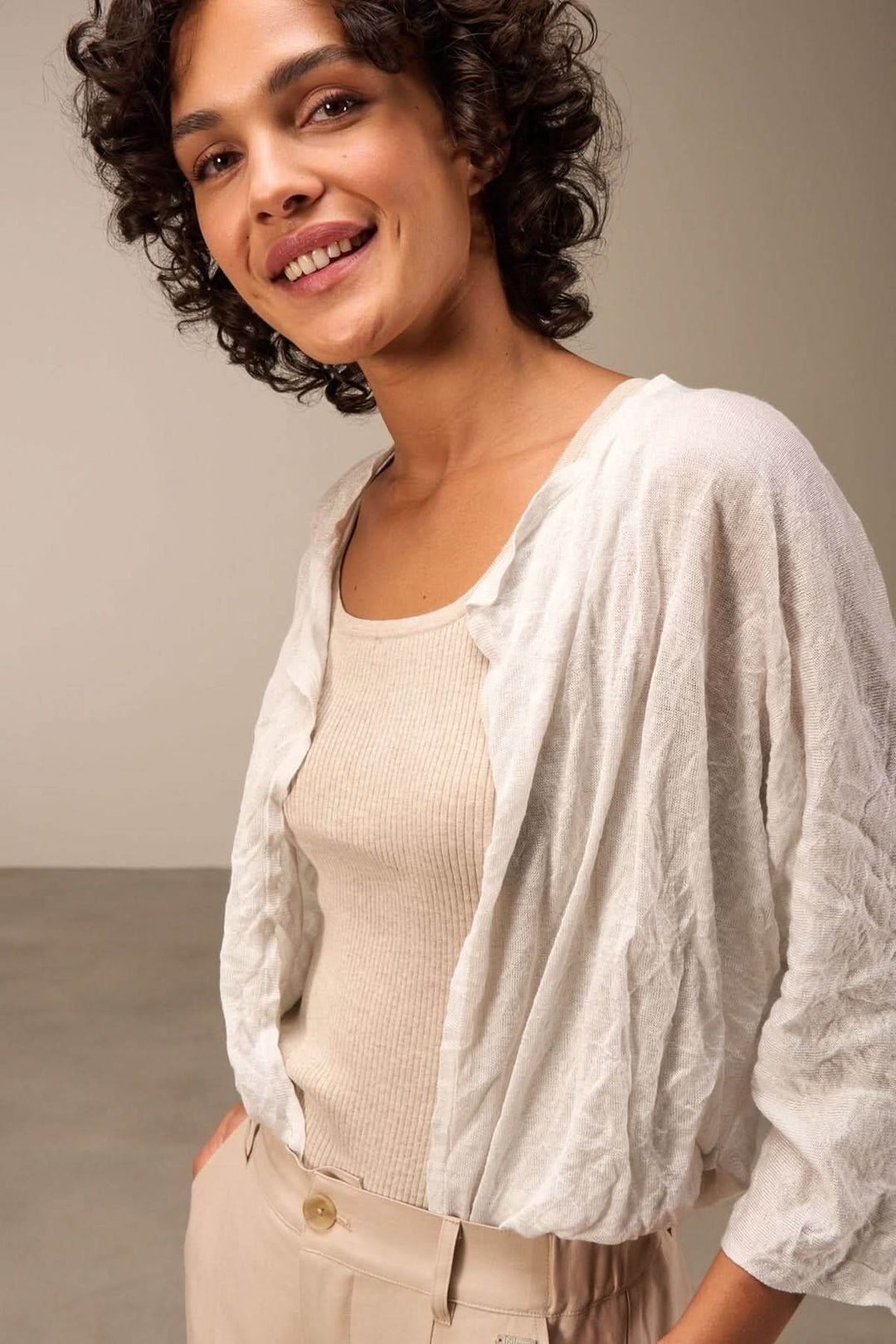 Femme souriante portant une veste en tissu léger blanche, avec un débardeur beige et un pantalon fluide.
