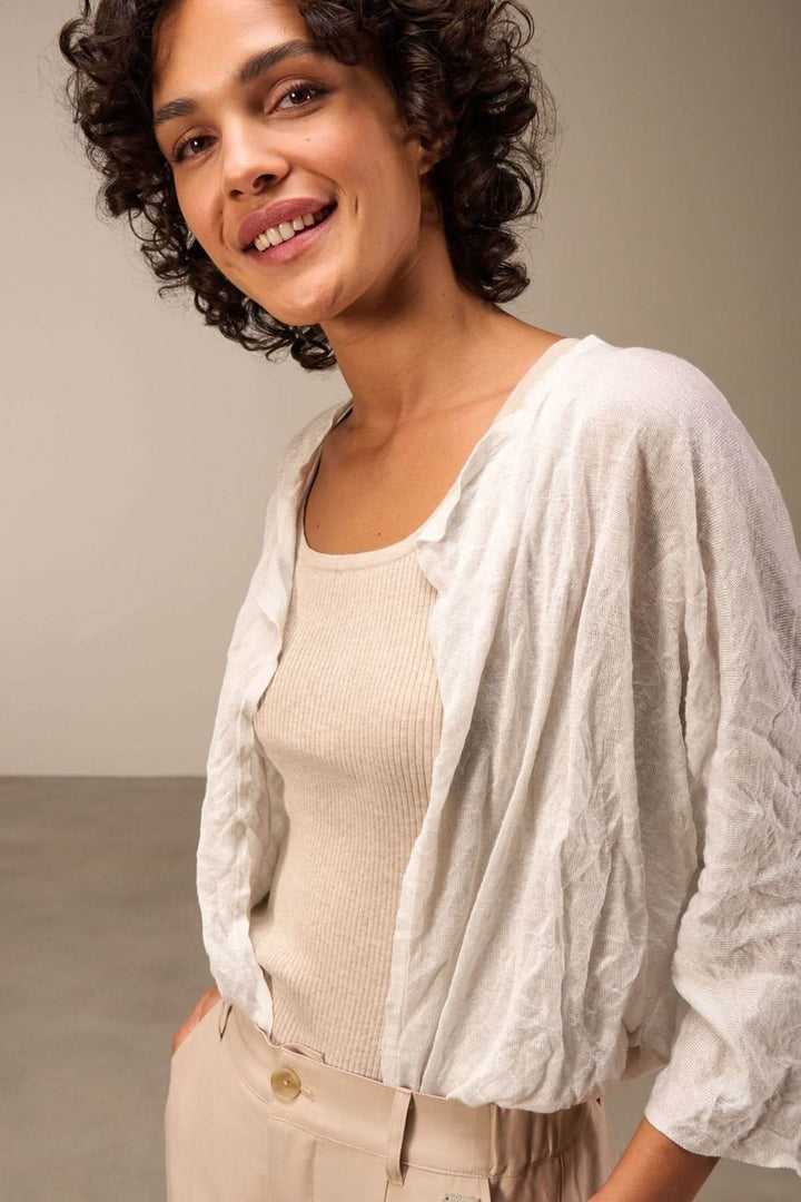 Femme souriante portant une veste en tissu léger blanche, avec un débardeur beige et un pantalon fluide.