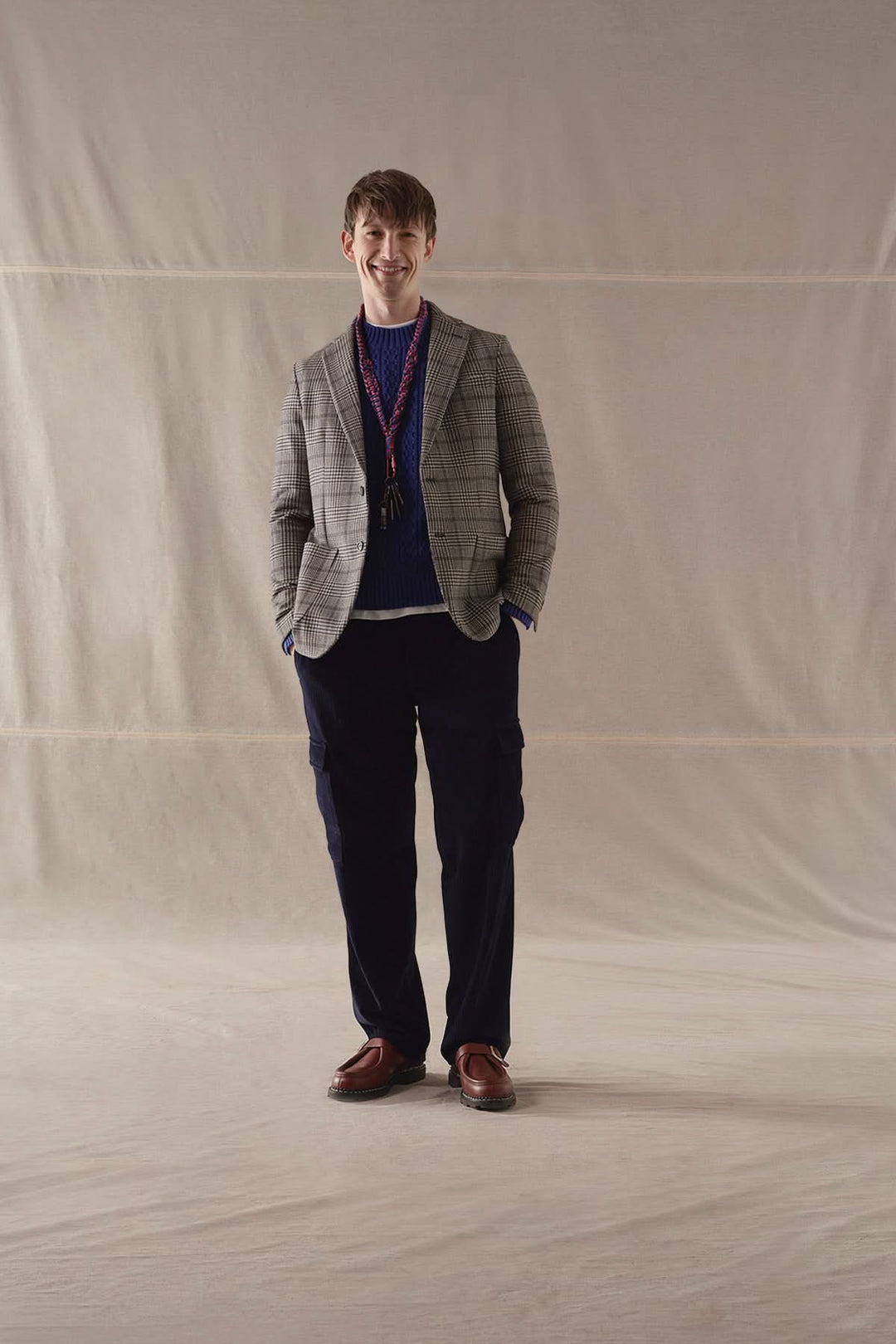 Homme souriant portant un veston à carreaux gris, un pull bleu et un pantalon sombre, sur fond neutre.