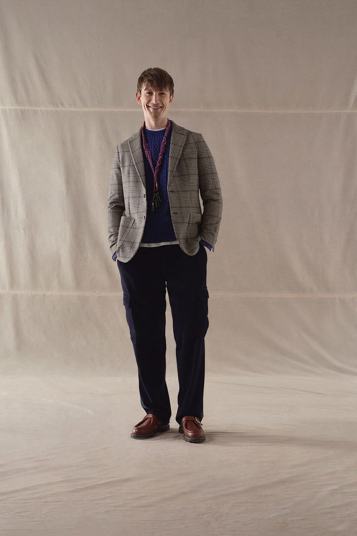 Homme souriant portant un veston à carreaux gris, un pull bleu et un pantalon sombre, sur fond neutre.