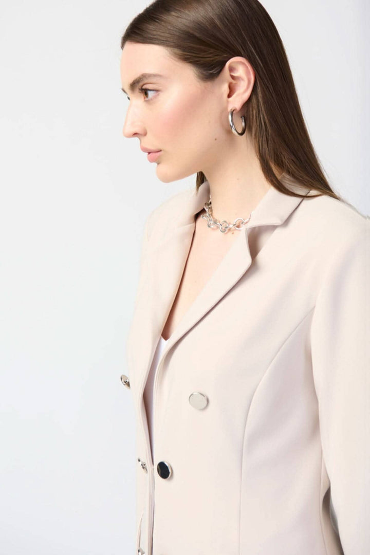 Femme en veston long beige, col cranté, boutons, et bijoux délicats, offrant un style élégant et contemporain.