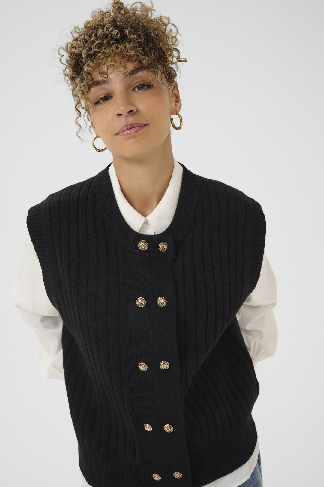 Veste Eroka sans manches en tricot à boutons dorés chic et polyvalente sur une chemise blanche, élégance décontractée.