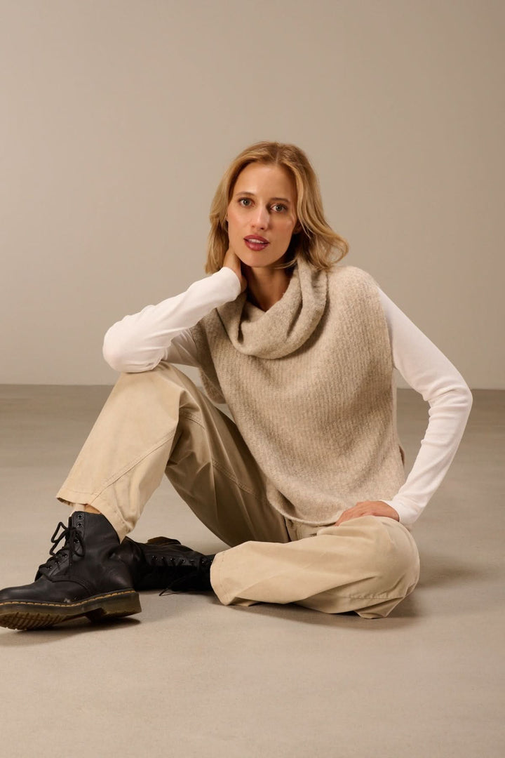 Femme portant un gilet sans manches en maille bouclée à col drapé, assise sur le sol avec un t-shirt à manches longues et pantalon beige.